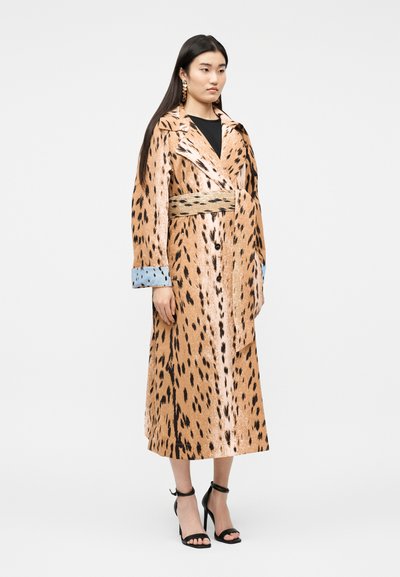 Γυναίκα που στέκεται φορώντας ένα μακρύ, καφέ παλτό με μαύρες κηλίδες και μπλε μανσέτες, μαύρο τοπ, χρυσά σκουλαρίκια και μαύρα σανδάλια με ψηλό τακούνι.