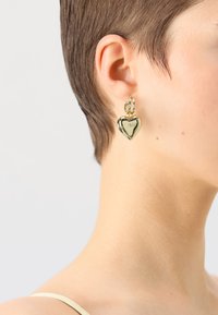 Gullhjerteformede øredobber med en polert finish, som har en liten løkke for festing. Glatt tekstur med et minimalistisk design.