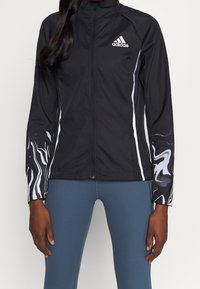 Musta Adidas-takki, jossa on kokovetoketju, valkoiset yksityiskohdat ja marmorin kuvioidut hihat. Kevyt, istuva malli, joka sopii aktiiviseen käyttöön.