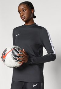 Μαύρη αθλητική μπλούζα με μακριά μανίκια και λευκό λογότυπο Nike με πλευρικές λεπτομέρειες. Το μοντέλο κρατά μια σχεδιασμένη ποδοσφαιρική μπάλα σε μαύρο και άσπρο χρώμα με κόκκινες λεπτομέρειες.