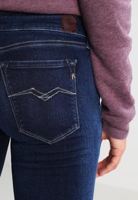 Vaqueros de denim azul oscuro con un diseño ajustado, que presentan un bolsillo trasero con un patrón de costura en zigzag y una etiqueta de parche de cuero en la cintura.