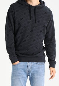 Osoba v černé mikině Adidas se vzorem opakujícího se loga a světle modrými džíny, s rukou v kapse, stojící na bílém pozadí.