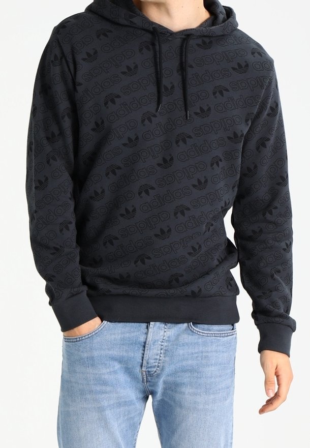 Osoba v černé mikině Adidas se vzorem opakujícího se loga a světle modrými džíny, s rukou v kapse, stojící na bílém pozadí.