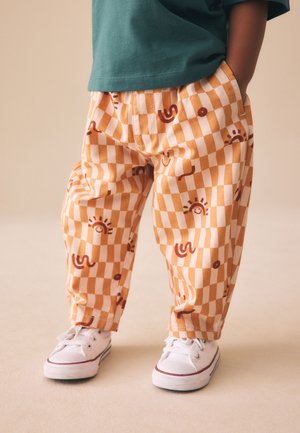 Niño con pantalones a cuadros naranja y blanco con patrones abstractos de sol y líneas onduladas, camiseta verde y zapatillas blancas, de pie con las manos en los bolsillos.
