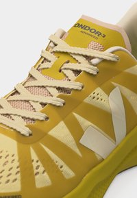 Κίτρινο και μπεζ αθλητικό παπούτσι με υφασμένα κορδόνια, πάνω μέρος από δίχτυ και ετικέτα "CONDOR 3 ADVANCED" στη γλώσσα.