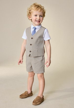 Băiat tânăr cu păr blond și creț stă zâmbind, purtând vestă și pantaloni scurți în carouri, cămașă albă, cravată albastră și pantofi maro slip-on.