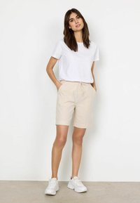 T-shirt blanc à manches courtes associé à un short à cordon de serrage beige. Le modèle porte des chaussures de sport blanches. Un design simple et des textures de tissu légères sont visibles.