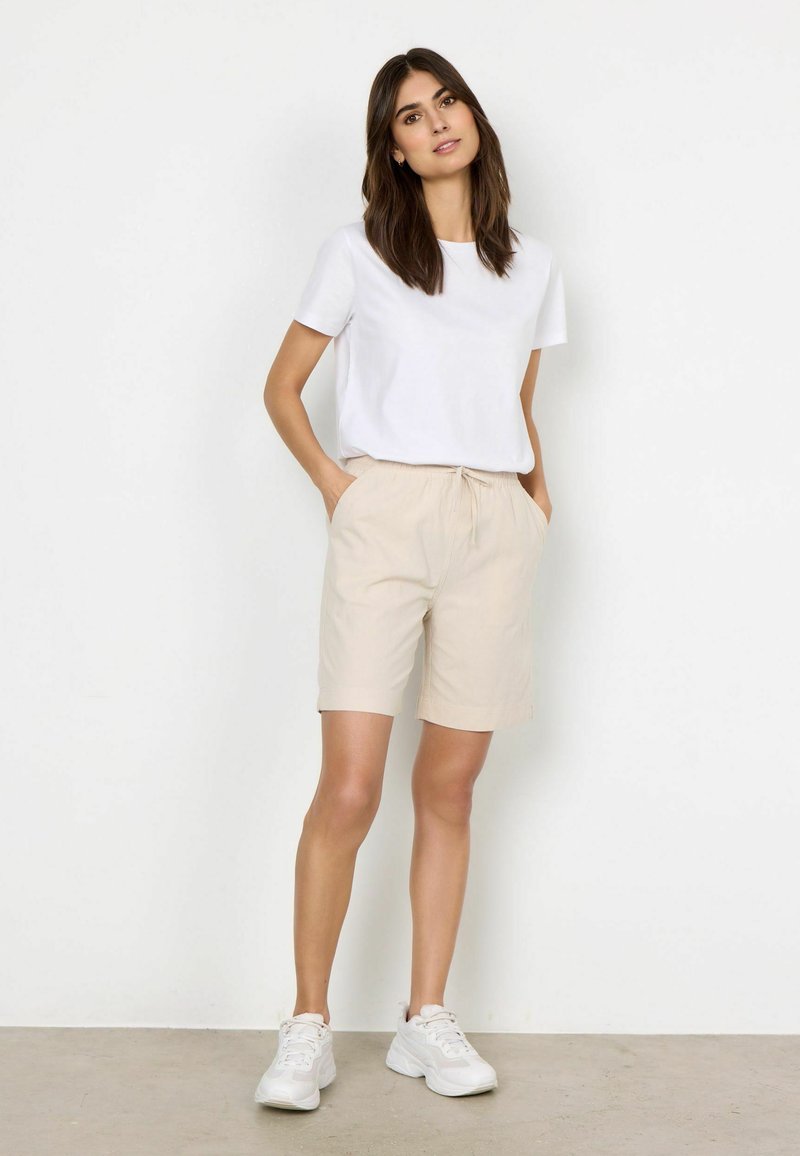 T-shirt blanc à manches courtes associé à un short à cordon de serrage beige. Le modèle porte des chaussures de sport blanches. Un design simple et des textures de tissu légères sont visibles.