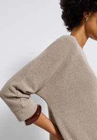 Beżowy sweter z dzianiny o luźnym kroju, z długimi rękawami i fakturą prążkowaną. Na nadgarstku modelki widnieje błyszczący brązowy bransoletka.