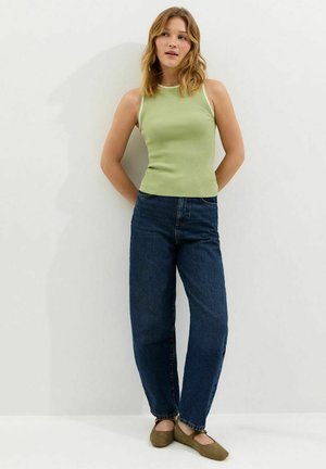 Donna in piedi contro una parete bianca che indossa una canottiera verde chiaro, jeans blu scuro a vita alta e scarpe piatte marroni, con le mani dietro la schiena.