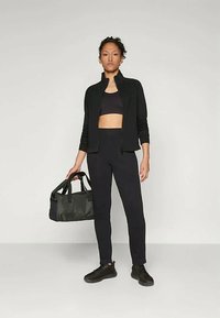 Svart träningskläder bestående av en zip-up jacka, en crop top och figurnära leggings. Modellen håller en svart gymväska och har på sig svarta sneakers.