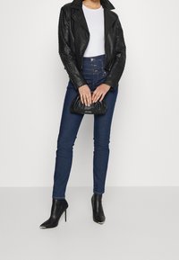 Chaqueta de moto de cuero negro con mangas con tachuelas, camiseta blanca, jeans azul de cintura alta, clutch negro y botines negros de tacón alto.