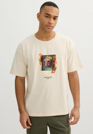 Joven con camiseta beige con un gráfico de una foto en llamas y el texto "Upper East Side NYC", de pie frente a un fondo claro y sencillo.