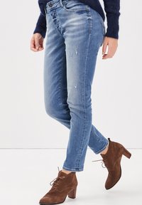 Jeans de mezclilla azul ajustados con detalles de desgaste y desgarros, combinados con botas cortas de ante marrón con tacón cuadrado.