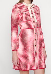 Czerwona i biała sukienka w wzór houndstooth z długimi rękawami, wykończona kokardą, z kieszeniami z przodu i złotymi guzikami wzdłuż środka. Teksturowany materiał.
