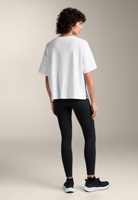 Vit oversized t-shirt med nedhängda ärmar, sidoklyftor och en mjuk textur, kombinerad med svarta leggings och svarta sneakers.