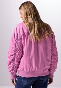 Różowa kurtka bombowiec z prążkowanymi mankietami i dołem, z bufiastymi rękawami i gładką, błyszczącą fakturą. Noszona na niebieskich dżinsach.