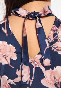 Blusa floreale navy con scollatura a V, texture setosa e dettaglio di nastro legato sul retro, con motivi di fiori rosa e bianchi.