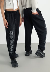 Black sweatpants featuring white graphic text on the left leg, with an elastic waistband and side pockets. Textured fabric with a loose fit.