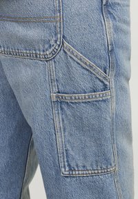 Pantalons cargo en denim bleu clair avec une grande poche latérale, des coutures visibles et une texture légèrement décolorée.
