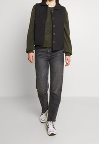 Gilet trapuntato nero con colletto, indossato sopra un maglione verde. Abbinato a jeans scuri e sneakers bianche con dettagli arancioni.