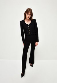 Černý zkrácený blazer se zlatými knoflíky a předními kapsami, zkombinovaný s přiléhavými černými kalhotami s rozšířenými nohavicemi a špičatými černými podpatky. Hladká textura.