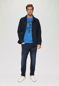 Niebieska bluza z czarnym napisem pod ciemną kurtką, w połączeniu z ciemnymi dżinsami i białymi sneakersami. Gładki materiał i swobodny krój.