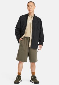 Zwarte lichtgewicht jas over een beige overhemden met knopen, gecombineerd met olijfgroene shorts en zwarte wandelschoenen. Gestructureerde stoffen en een relaxte pasvorm.