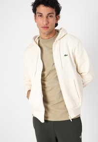 Lys beige zip-up hættetrøje med en forkantlomme, parret med en lys olivengrøn T-shirt. Har et lille grønt krokodillelogo på brystet.