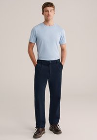 Világoskék pamut póló tengerészkék nadrággal párosítva. A nadrág gombos záródással és zsebekkel rendelkezik. A modell a zsebeiben tartja a kezét.