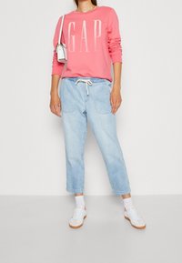 Rosa sweatshirt med "GAP" logotyp, ljusblå avslappnade denimbyxor, vita sneakers och liten vit axelväska. Avslappnad outfit på modell.