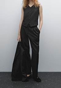 Donna che indossa un gilet nero senza maniche con bottoni e pantaloni a gamba larga, che tiene un giubbotto nero, mentre è in piedi contro un muro semplice su un pavimento maculato.