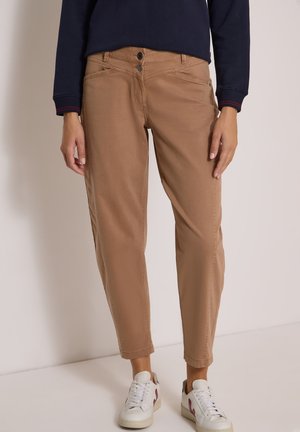 Femme portant un pantalon marron clair avec deux boutons devant et des baskets blanches avec des accents roses, debout devant un fond uni.