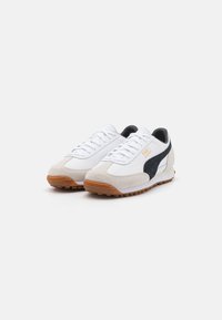 Zapatillas blancas con parte superior de ante y textil, franja lateral negra y suela de goma. Cuenta con un diseño clásico de cordones y puntera redondeada.