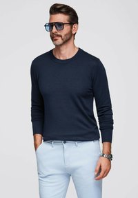 Marineblauer Pullover mit Rundhalsausschnitt, enganliegenden Ärmeln, kombiniert mit hellblauen Hosen. Die Accessoires umfassen große dunkle Sonnenbrillen und eine silberne Uhr.