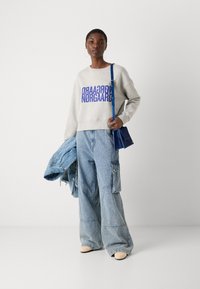 Grå hettegenser med lilla tekst, lyseblå cargo-jeans og beige sko. Holder en blå håndveske og har en denimjakke over skuldrene.