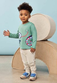 Lichtgroene langmouwen shirt met een kleurrijke slakafbeelding, beige sweatpants en blauwe slippers, staande naast een getextureerde ronde kruk.