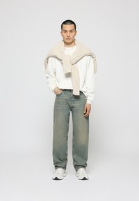 White long-sleeve shirt, light beige knitted sweater draped over shoulders, blue loose-fit jeans, and white sneakers. Plain background.