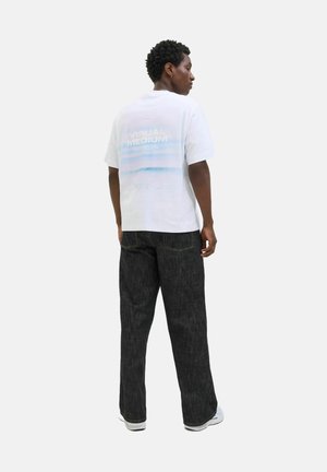 Witte katoenen t-shirt met een grafische opdruk "VISUAL MEDIUM" in pastelkleuren op de rug, gecombineerd met donkere spijkerbroek en witte sneakers.