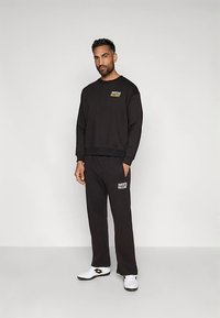 Άνδρας στέκεται μπροστά σε απλή τοιχογραφία, φορώντας μαύρη φούτερ και παντελόνι με λευκά λογότυπα, σε συνδυασμό με λευκά αθλητικά παπούτσια.