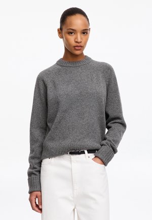 Femme aux cheveux tirés en arrière portant un pull en maille gris rentré dans un pantalon blanc avec une ceinture noire, debout avec une main dans la poche.