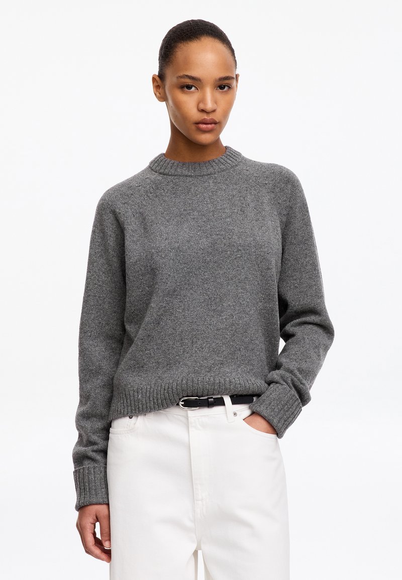 Femme aux cheveux tirés en arrière portant un pull en maille gris rentré dans un pantalon blanc avec une ceinture noire, debout avec une main dans la poche.
