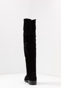 Bota de ante negra sobre la rodilla con un pequeño tacón, que cuenta con un zipper vertical y una hebilla decorativa en la parte superior. Textura suave y mate.