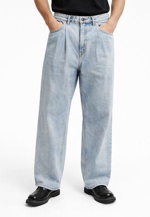 Person trägt hellblaue Jeans mit weitem Bein und schwarze Lederschuhe, Hände in den vorderen Taschen, steht vor weißem Hintergrund.