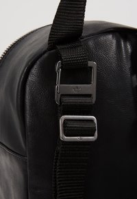 Black leather bag with a textured surface, featuring a sturdy adjustable strap and metal hardware with an insignia. Zipper closure detail visible.
