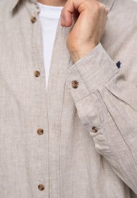 Camisa de botones a rayas beige con una textura suave, que presenta botones marrones y un pequeño logo en el pecho. Mangas largas dobladas en el puño.