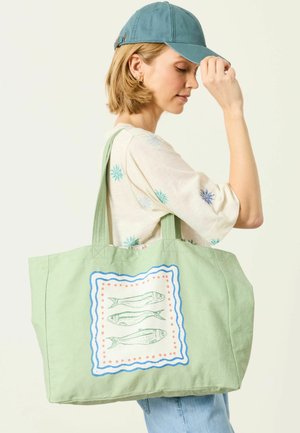 Nainen sinisessä lippalakissa ja kukallisessa paidassa kantaa isoa vaaleanvihreää kangaskassia, jossa on kalan kuva sivulla.