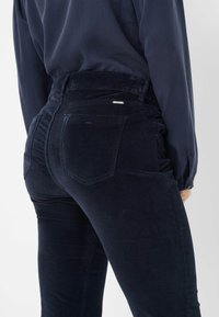 Jeans taille haute en velours côtelé bleu marine avec une silhouette élégante, dotés de poches arrière et d'un subtil détail métallique au niveau de la ceinture.