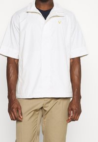 Mężczyzna w białej kurtce zapinanej na zamek błyskawiczny z krótkim rękawem i żółtym logo ptaka oraz beżowych spodniach, stojący na tle jednolitym.