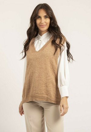 Gilet en tricot beige avec un design en V, porté sur une blouse blanche à manches bouffantes. Texture lisse, ourlet côtelé et coupe décontractée.